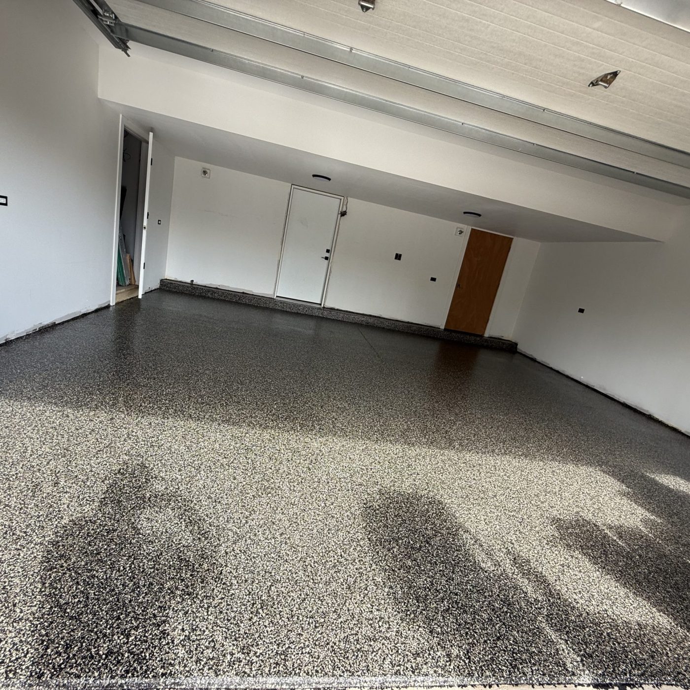 This image shows a freshly finished black and gray epoxy-coated garage floor. The surface has a glossy sheen, reflecting light from the open garage door and creating a clean, polished look. The epoxy has a speckled appearance, with a mix of dark and light flakes that give the floor a textured, high-end finish. The overall result looks professional and durable, giving the garage a modern and refined appearance.